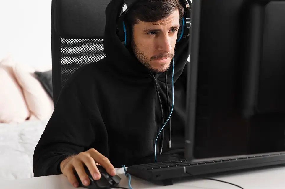 Focused young man in a black hoodie and blue headphones looking at a computer monitor while holding a mouse