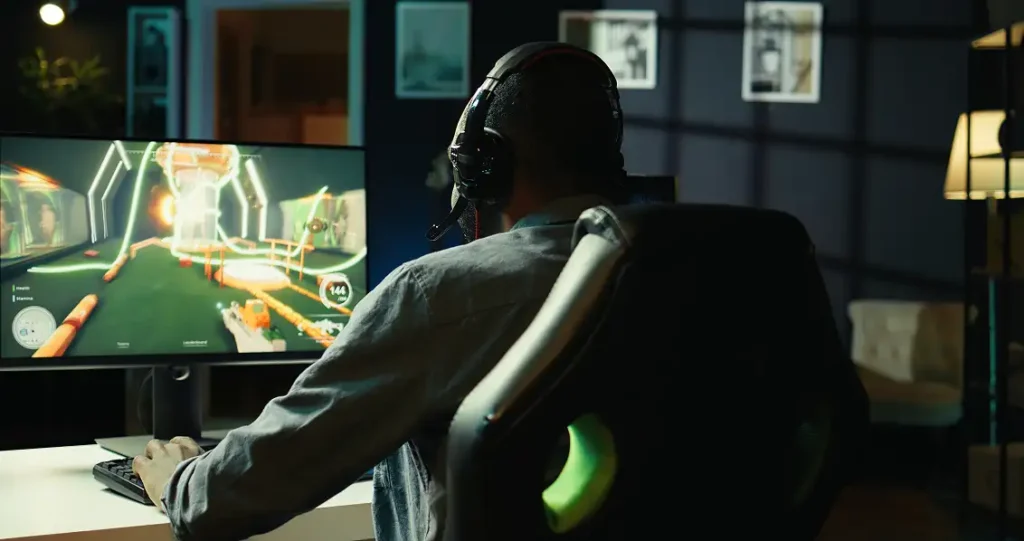Rear view of a man wearing a headset sitting in a gaming chair and playing a first-person shooter game on a monitor