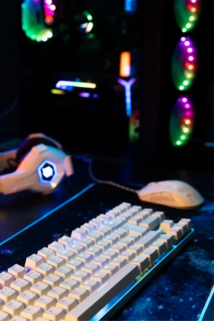 Close-up shot of a white mechanical gaming keyboard with RGB illuminated computer tower and gaming headset in a dark room